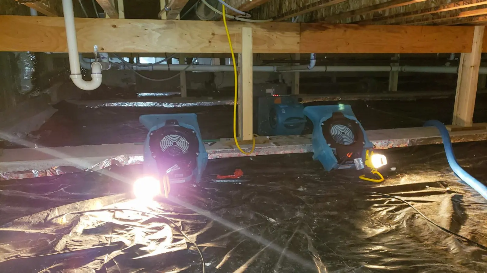 Crawl space with plastic sheeting, air movers and a work light under the floor joists